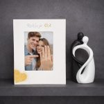 Bijeli drveni okvir za zaruke s rukom ispisanim imenima zaručnika i datumom, ukrašen motivima prstenja i srca.